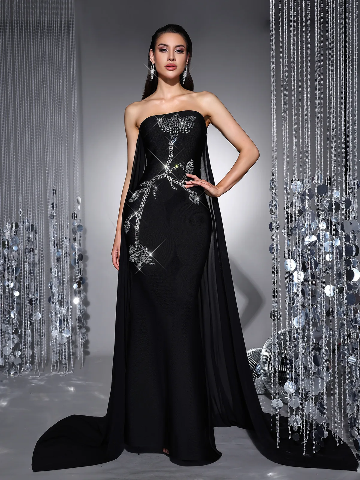 Model wearing black strapless bandage evening gown with rhinestone embroidery and mesh drape panels, front view